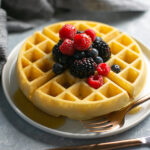 crispy waffle topped with mixed berries on white plate, fork and knife on the side